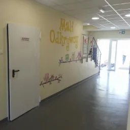 Korytarz w przedszkolu z pomalowaną ścianą, na której widnieje napis 'Mali Odkrywcy' i rysunki sów na gałęziach, białe drzwi po lewej stronie i widok na schody oraz wyjście na końcu korytarza.