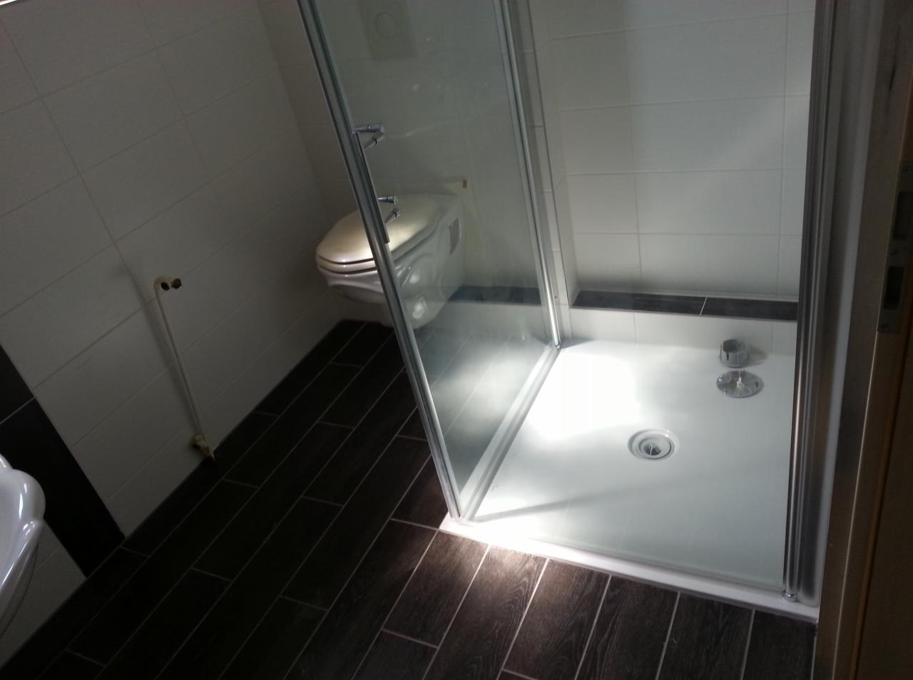 Kabina prysznicowa z brodzikiem, widok z góry, ciemna podłoga imitująca drewno, białe płytki na ścianach, toaleta w tle.