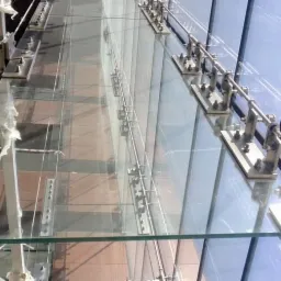 Szklana fasada mocowana punktowo do stalowej konstrukcji wsporczej, widoczne elementy mocujące i żebra szklane, częściowo zabezpieczone folią ochronną na czas montażu.