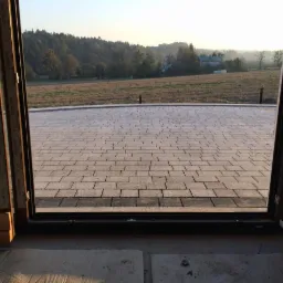 Widok z wnętrza przez otwarte drzwi balkonowe na nowo ułożoną kostkę brukową na zewnątrz, z polami i drzewami w tle. Kostka w odcieniach szarości i beżu.