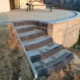 Ułożone schody z kostki brukowej prowadzące na okrągły taras. Widoczne ciemne obrzeża kostki i jasne wypełnienie. Tło stanowi budynek i zieleń.