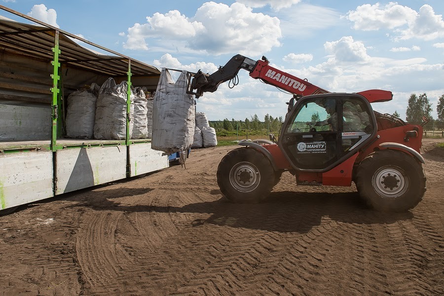 Czerwony wózek widłowy Manitou podnosi duży, szary worek z ładowni ciężarówki wypełnionej workami, na tle piaszczystego terenu i błękitnego nieba z chmurami.