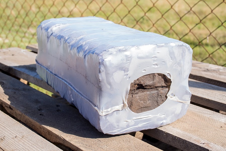 Paczka brykietu opałowego owinięta w przezroczystą folię, z widocznym fragmentem brykietu przez otwór, leżąca na drewnianej palecie, w tle siatka ogrodzeniowa.