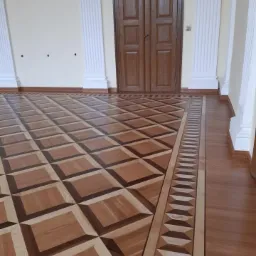 Elegancka podłoga parkietowa z geometrycznym wzorem w odcieniach brązu i beżu, ułożona w przestronnym wnętrzu z kolumnami i drewnianymi drzwiami.