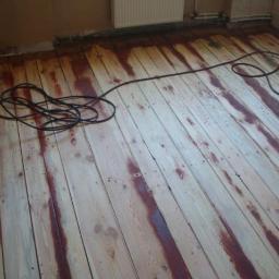 P.H.U PARKIET SZYMON STYŚ - Podłoga z desek w trakcie renowacji, widoczne ślady starej bejcy i częściowo usunięta warstwa wierzchnia, obok stoi maszyna do szlifowania.