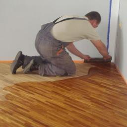 P.H.U PARKIET SZYMON STYŚ - Pracownik w kombinezonie aplikuje lakier na drewnianą podłogę w jodełkę przy krawędzi ściany, zabezpieczonej taśmą malarską.