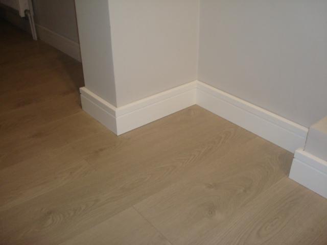 Jasne panele podłogowe ułożone w rogu pomieszczenia, z białymi listwami przypodłogowymi.