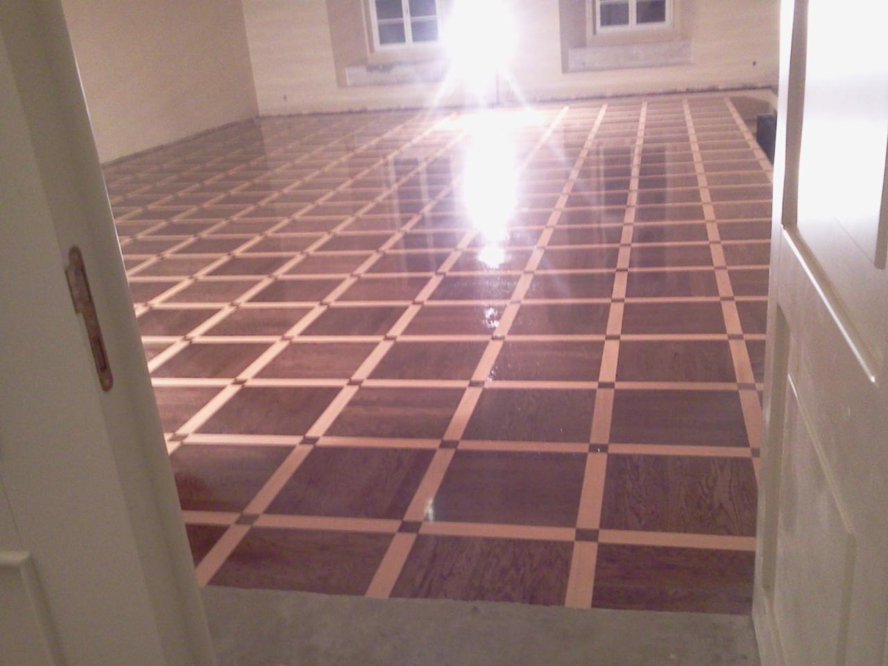 Nowo ułożona, błyszcząca podłoga parkietowa w kwadratowy wzór, widoczna zza uchylonych białych drzwi, z refleksami światła odbijającymi się od powierzchni.