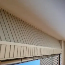 Fragment elewacji budynku z widoczną podbitką dachową, wykonaną z paneli PCV w kolorze beżowym, z elementami wentylacyjnymi i listwami ozdobnymi, widziany z dołu pod kątem.