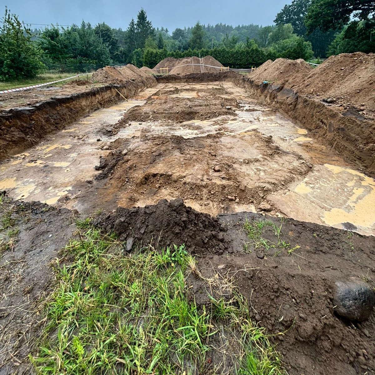 Wykonany wykop pod fundamenty domu, widoczne ślady wody i błota. W tle usypane pryzmy ziemi i zieleń drzew. Ujęcie z bliska na trawę i ziemię.