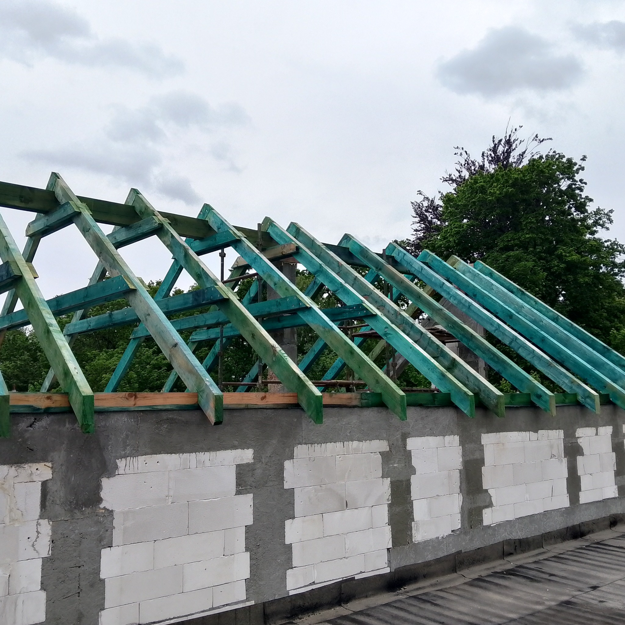 Wznoszenie konstrukcji dachu z impregnowanego drewna na murze z betonu komórkowego. Widoczne fragmenty drzew na tle pochmurnego nieba.