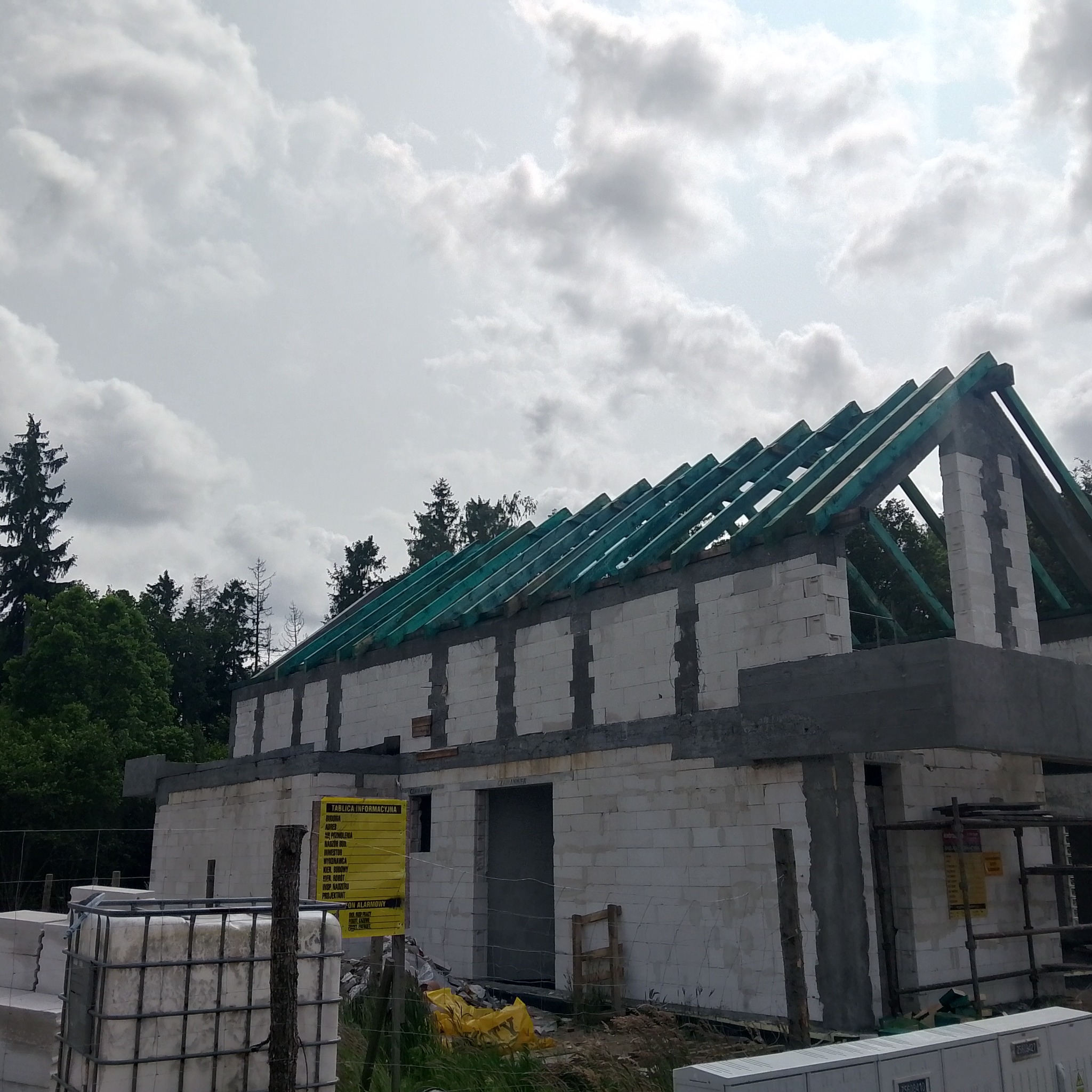 Budowa domu z białych bloczków, widoczne zielone krokwie dachu i tablica informacyjna. Stan surowy otwarty w słoneczny dzień z chmurami w tle.