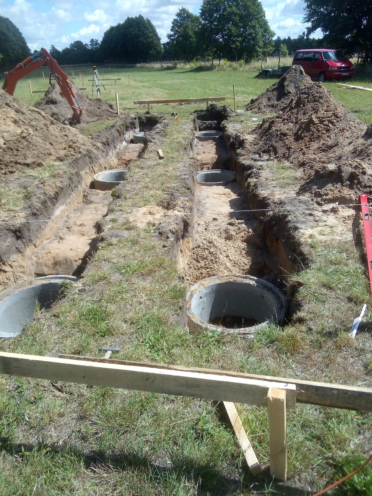 Wykopane rowy pod fundamenty z ustawionymi betonowymi kręgami. Na trawiastym terenie widoczna koparka i poziomica. Prace ziemne w słoneczny dzień.