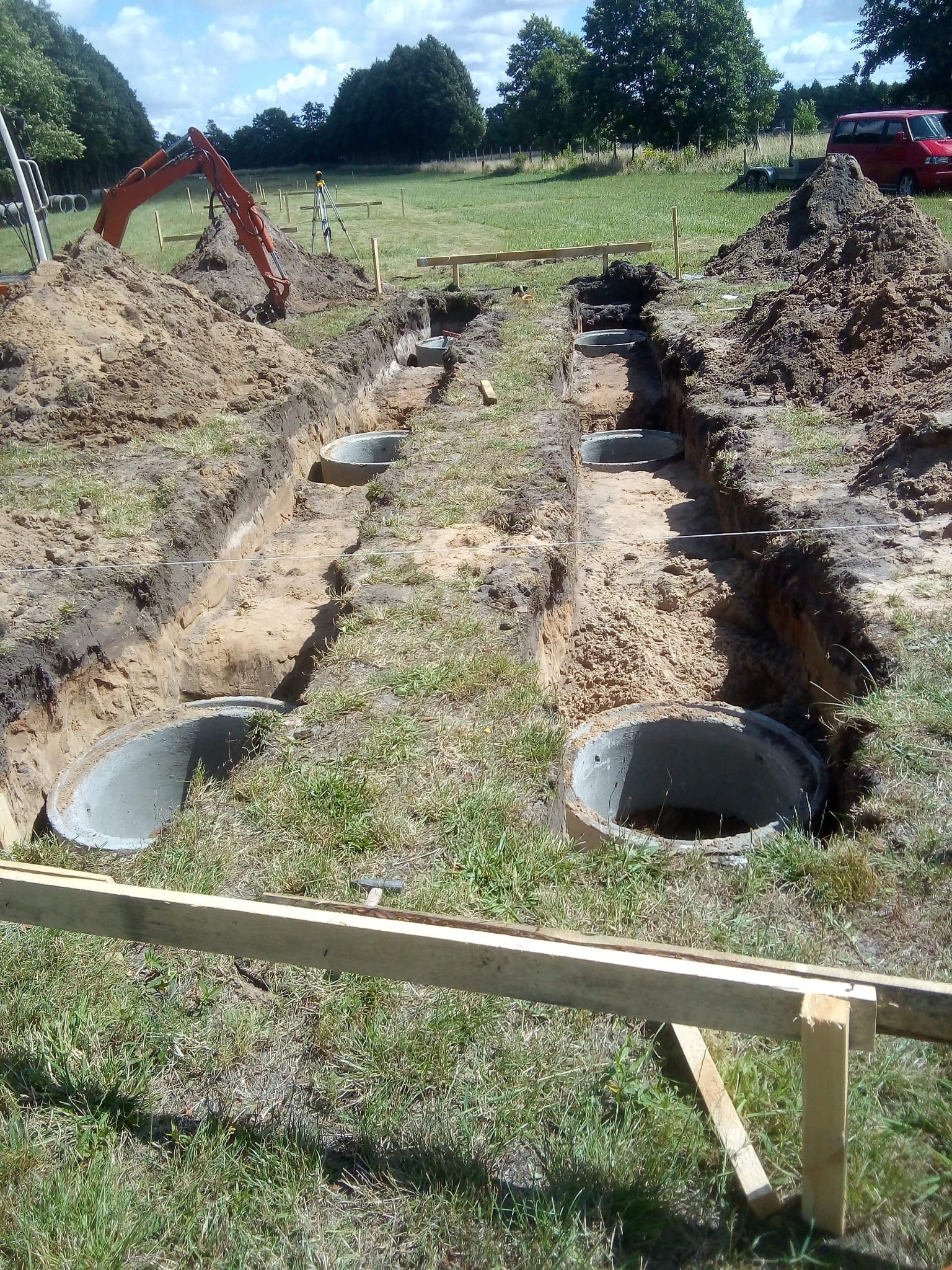 Wykopane rowy pod fundamenty z betonowymi kręgami w ziemi, widoczny sprzęt budowlany i ułożone drewniane szalunki. Prace ziemne w toku, słoneczny dzień.