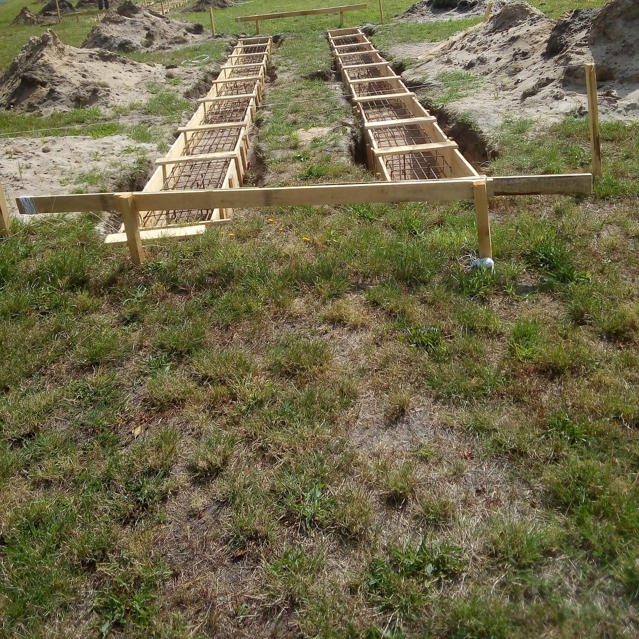 Zbrojenie i szalunek w wykopie pod fundament. Dwa rzędy przygotowane do zalania betonem, widoczne drewniane elementy konstrukcji.