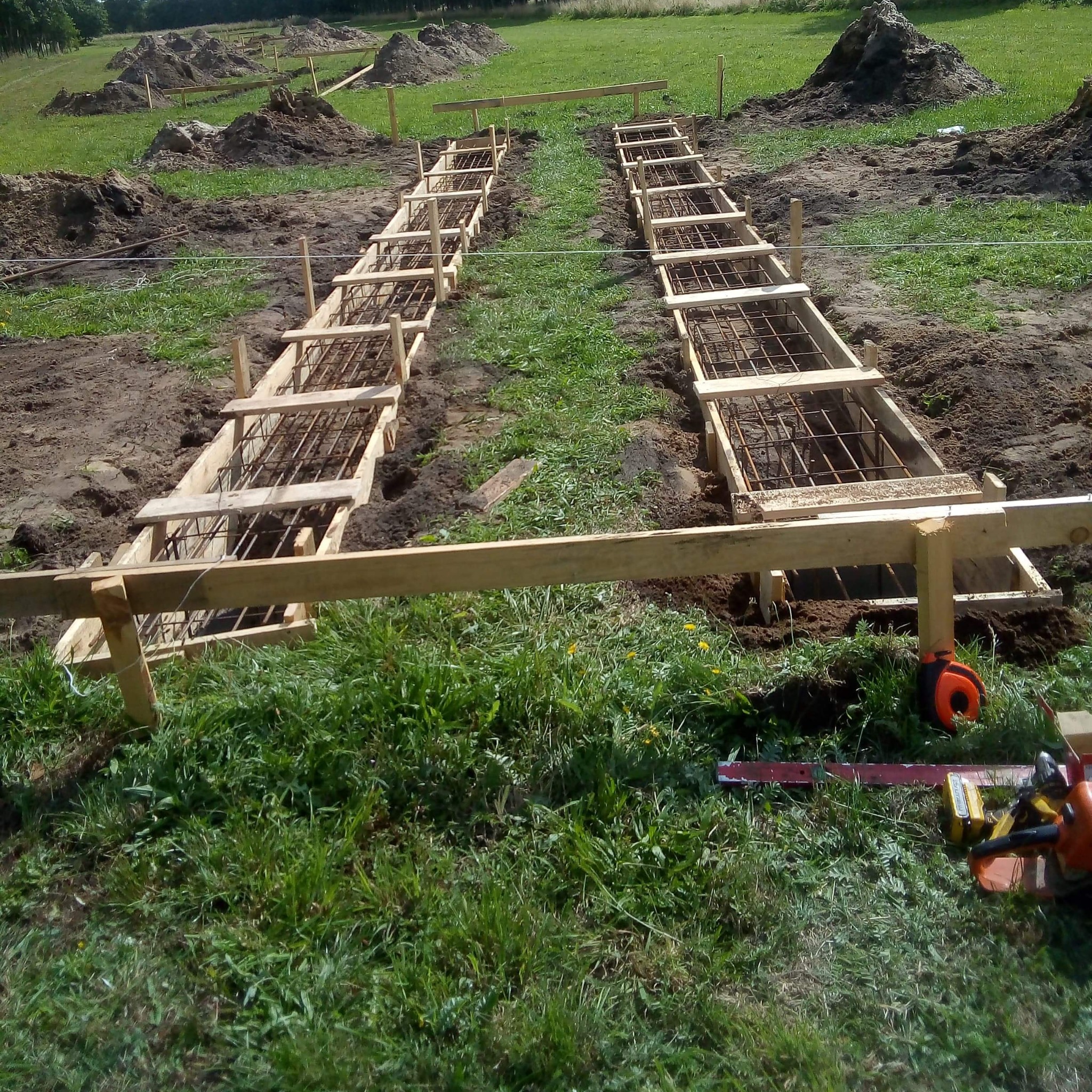 Przygotowanie szalunków i zbrojenia pod fundamenty na zielonej działce. Widoczne narzędzia: piła łańcuchowa i miara. W tle pryzmy ziemi.