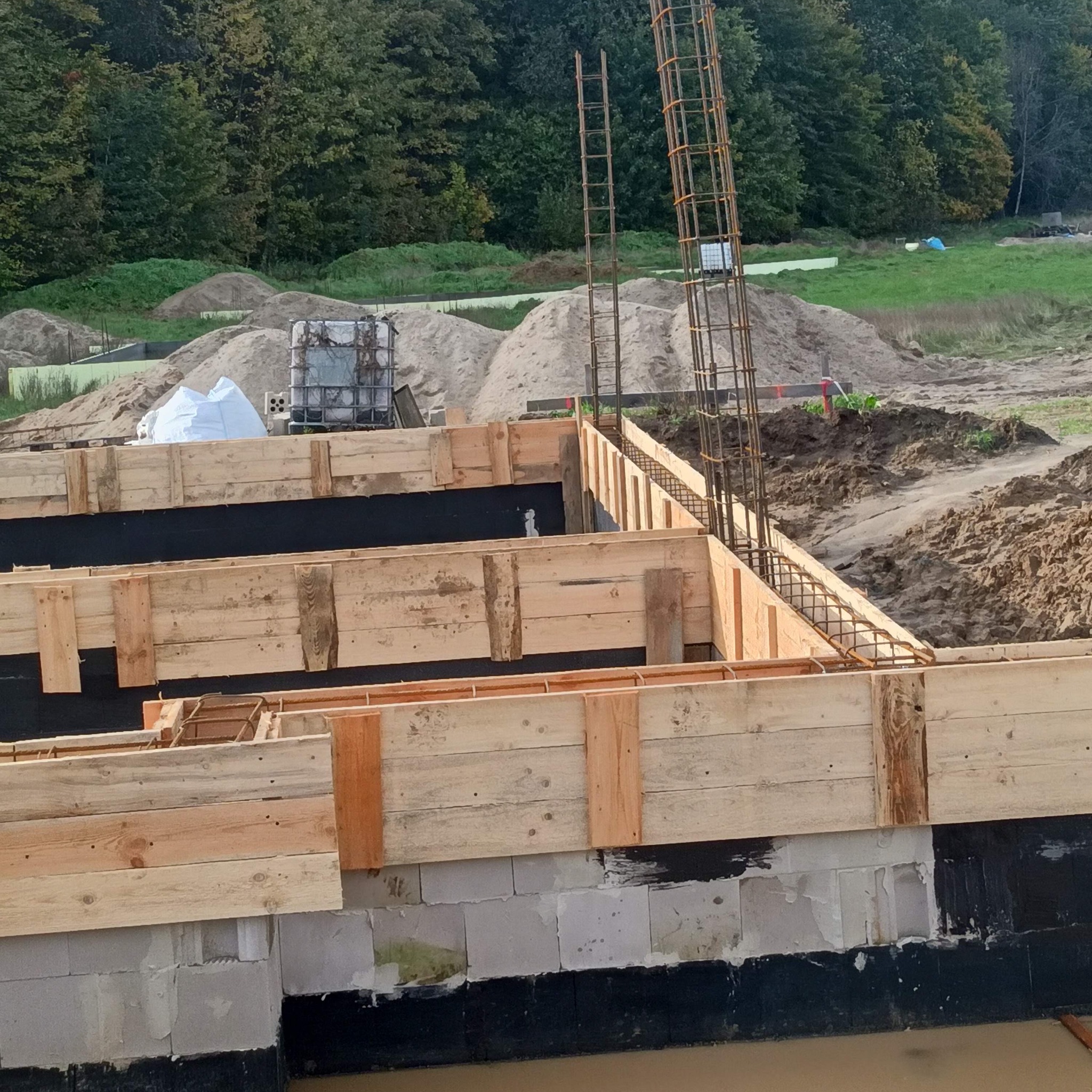 Szczegółowy widok szalunku fundamentów z wystającymi prętami zbrojeniowymi na tle zieleni i piasku, ukazujący etap budowy domu.