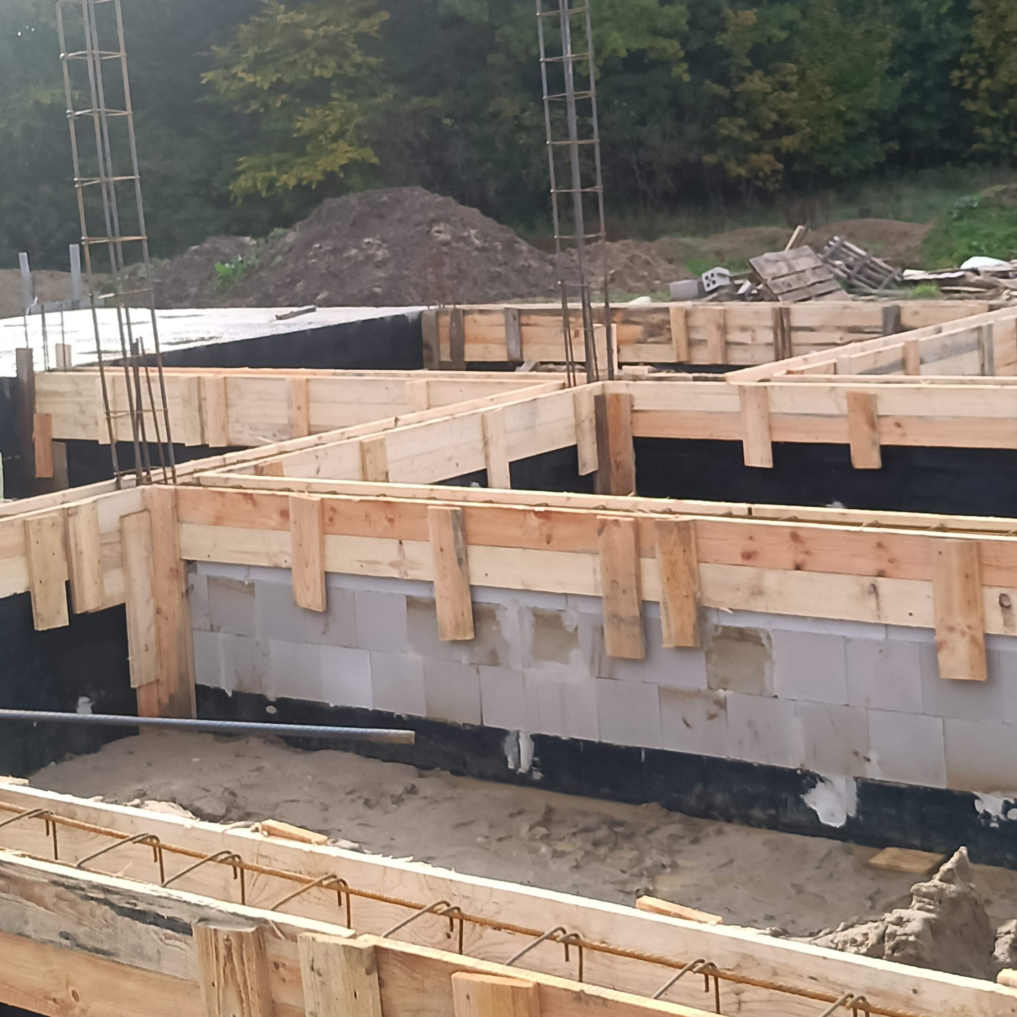 Zbrojone, drewniane szalunki fundamentów z izolacją pionową, widoczne pręty zbrojeniowe i bloczki fundamentowe, w tle ziemia i zieleń.