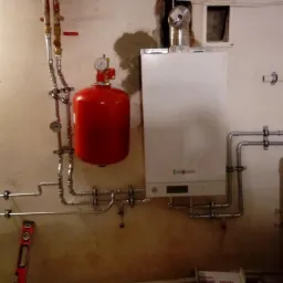 Instalacja kotła gazowego Viessmann z czerwonym zbiornikiem wyrównawczym, widoczne rury instalacji grzewczej i poziomica na podłodze, surowy stan wykończenia ściany.