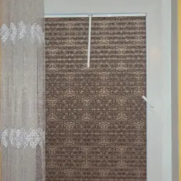 Okno z plisowaną roletą w brązowe wzory, zasłonięte częściowo firanką z delikatnym, białym wzorem