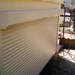 Zewnętrzna, beżowa roleta zamontowana na budynku, widoczna z boku, z fragmentem altany w tle.