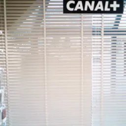 Beżowe żaluzje poziome w pomieszczeniu, na tle okna widoczny napis CANAL+.