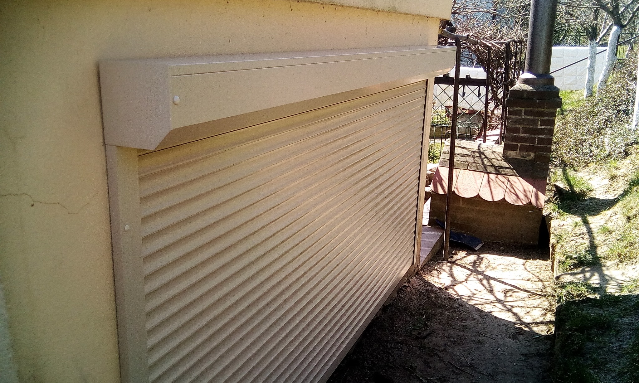 Zewnętrzna, beżowa roleta zamontowana na budynku, widoczna z boku, z fragmentem altany w tle.