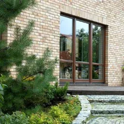 Nowoczesny dom z cegły z dużymi, brązowymi oknami, widok z ogrodu z kamienną ścieżką i sosną na pierwszym planie.