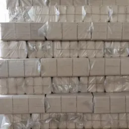 Paleta z ułożonymi warstwami brykietu drzewnego w szarym papierze i folii. Widoczne pęknięcia na podłodze. Część materiału poza kadrem.