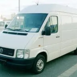 Biały bus dostawczy z wysokim dachem, zaparkowany na zewnątrz, widok z przodu pod lekkim kątem, szare tło parkingu. Widoczny fragment drugiego auta.