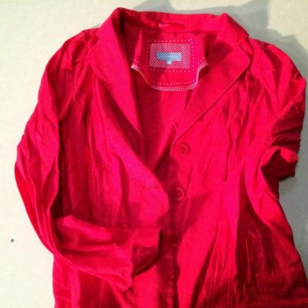 Czerwona, damska marynarka z krótkim rękawem, leżąca na jasnym tle. Widoczne dwa guziki i metka z delikatnym wzorem w kropki.