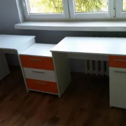 Białe biurko na wymiar z pomarańczowymi akcentami, ustawione pod oknem. Widoczne trzy szuflady i grzejnik. Prosta forma, jasne kolory, funkcjonalne rozwiązanie.