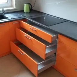 Nowoczesna kuchnia z pomarańczowymi frontami i wysuwanymi szufladami. Blat granitowy, białe szafki górne. Widoczny czajnik elektryczny.