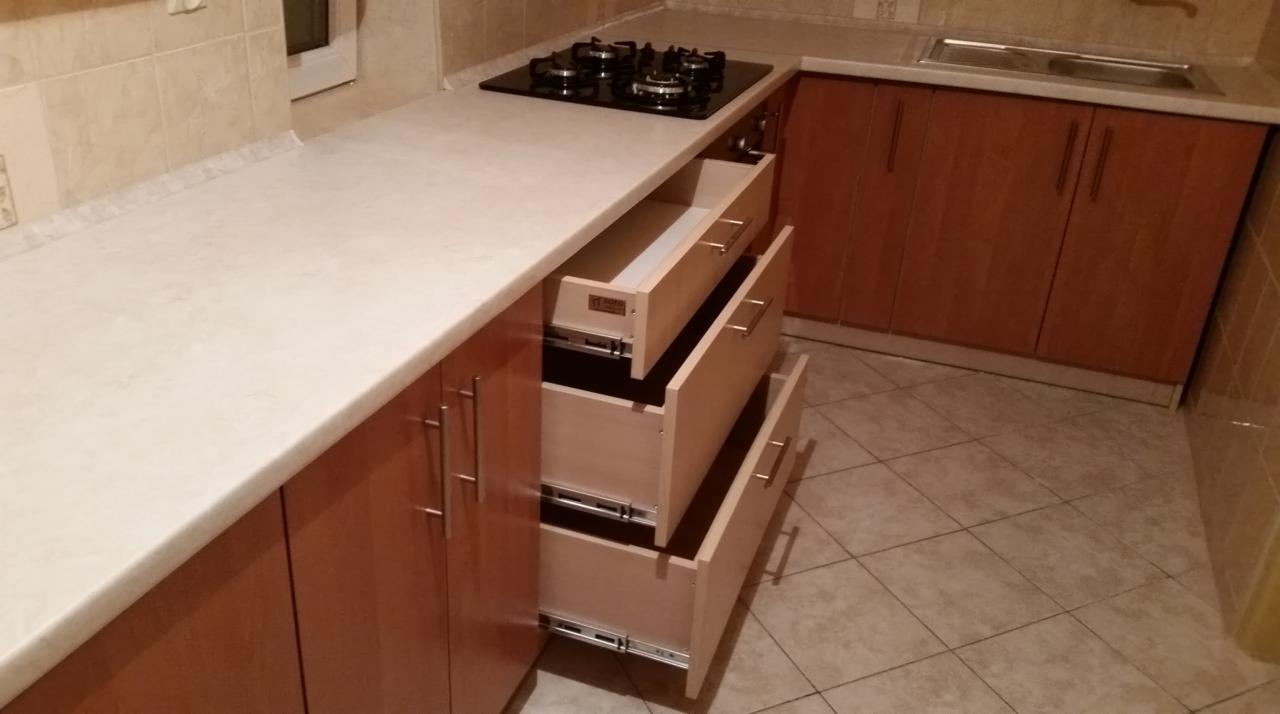 Nowoczesna kuchnia z drewnianymi szafkami i blatem, otwarte szuflady z widocznym systemem prowadnic, płyta grzewcza na blacie. Ujęcie z perspektywy podłogi.