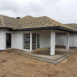 Nowy dom w trakcie budowy: białe ściany, okna PCV, przygotowana konstrukcja dachu z łatami i częściowo wykończony taras z betonowymi schodami.