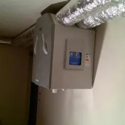 Szara jednostka centralna wentylacyjna z podłączonymi elastycznymi przewodami aluminiowymi, zamontowana w rogu pomieszczenia o surowym wystroju, widoczny fragment ściany i podłogi z drobnymi...