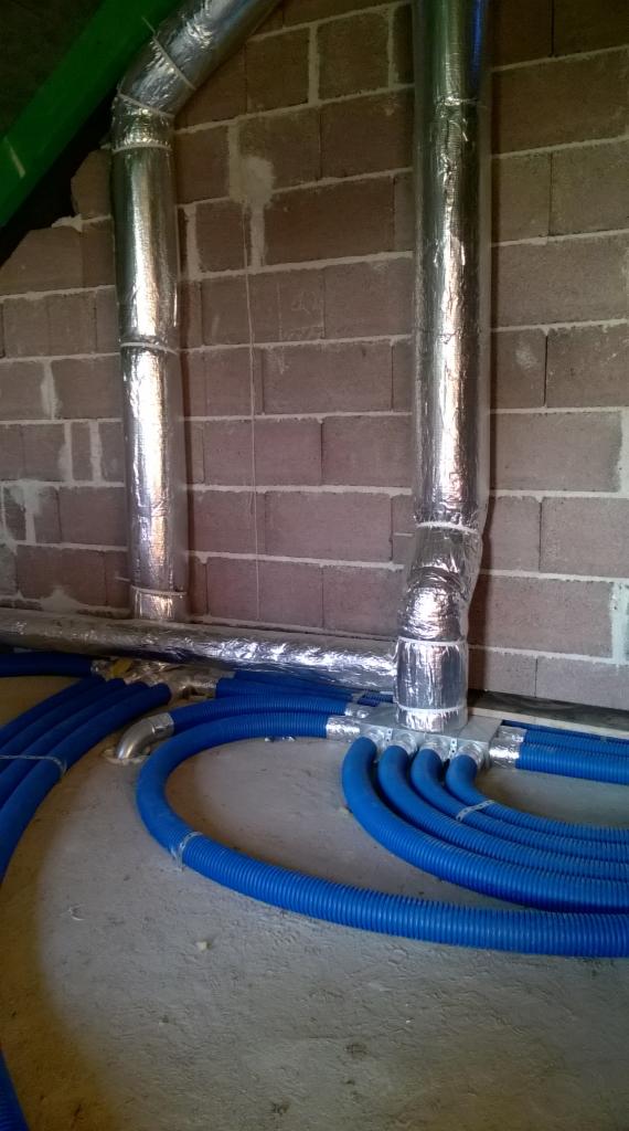 Instalacja wentylacyjna: srebrne, izolowane rury pionowe i niebieskie, karbowane przewody poziome na tle surowej ściany z bloczków.