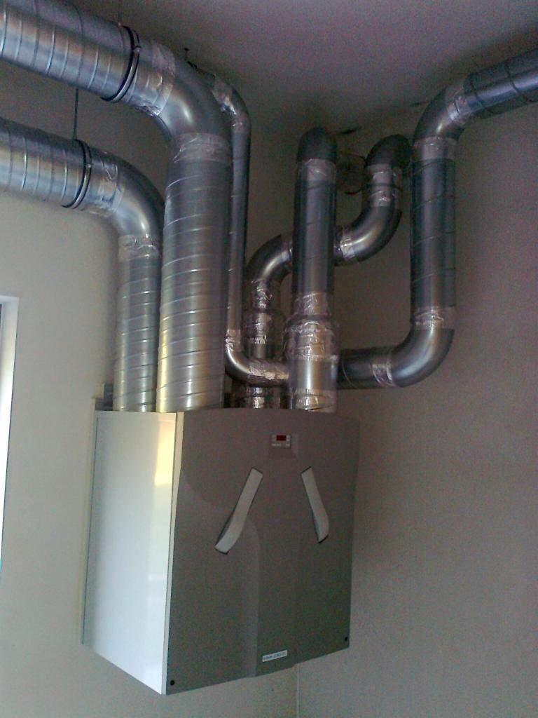 Szare urządzenie z podłączonymi, metalowymi rurami wentylacyjnymi w pomieszczeniu. Widoczne elementy instalacji i połączenia.
