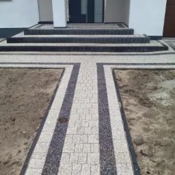 Nowoczesny chodnik z kostki brukowej w dwóch kolorach prowadzi do wejścia domu z trzema schodami. Widoczne studzienki kanalizacyjne.