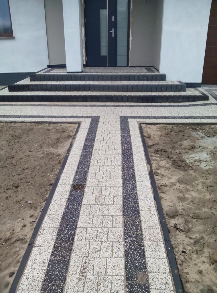 Nowoczesny chodnik z kostki brukowej w dwóch kolorach prowadzi do wejścia domu z trzema schodami. Widoczne studzienki kanalizacyjne.
