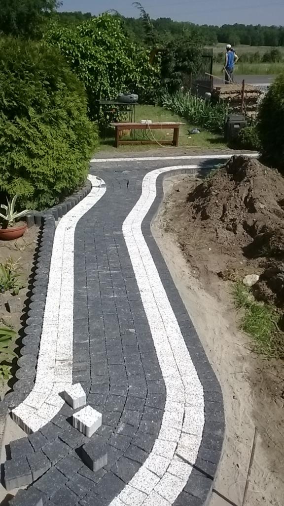 Kręta ścieżka z kostki brukowej w dwóch kolorach, otoczona zielenią i ziemią. Widoczne niedokończone fragmenty i narzędzia w tle, sugerujące trwający proces.