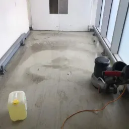 Podłoga w biurze w trakcie czyszczenia maszyną polerującą, widoczny kanister z płynem czyszczącym i mokre ślady po umytej powierzchni.