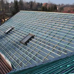 Dach w trakcie krycia: widoczne membrany, łaty i dwa okna dachowe, w tle zabudowa jednorodzinna.