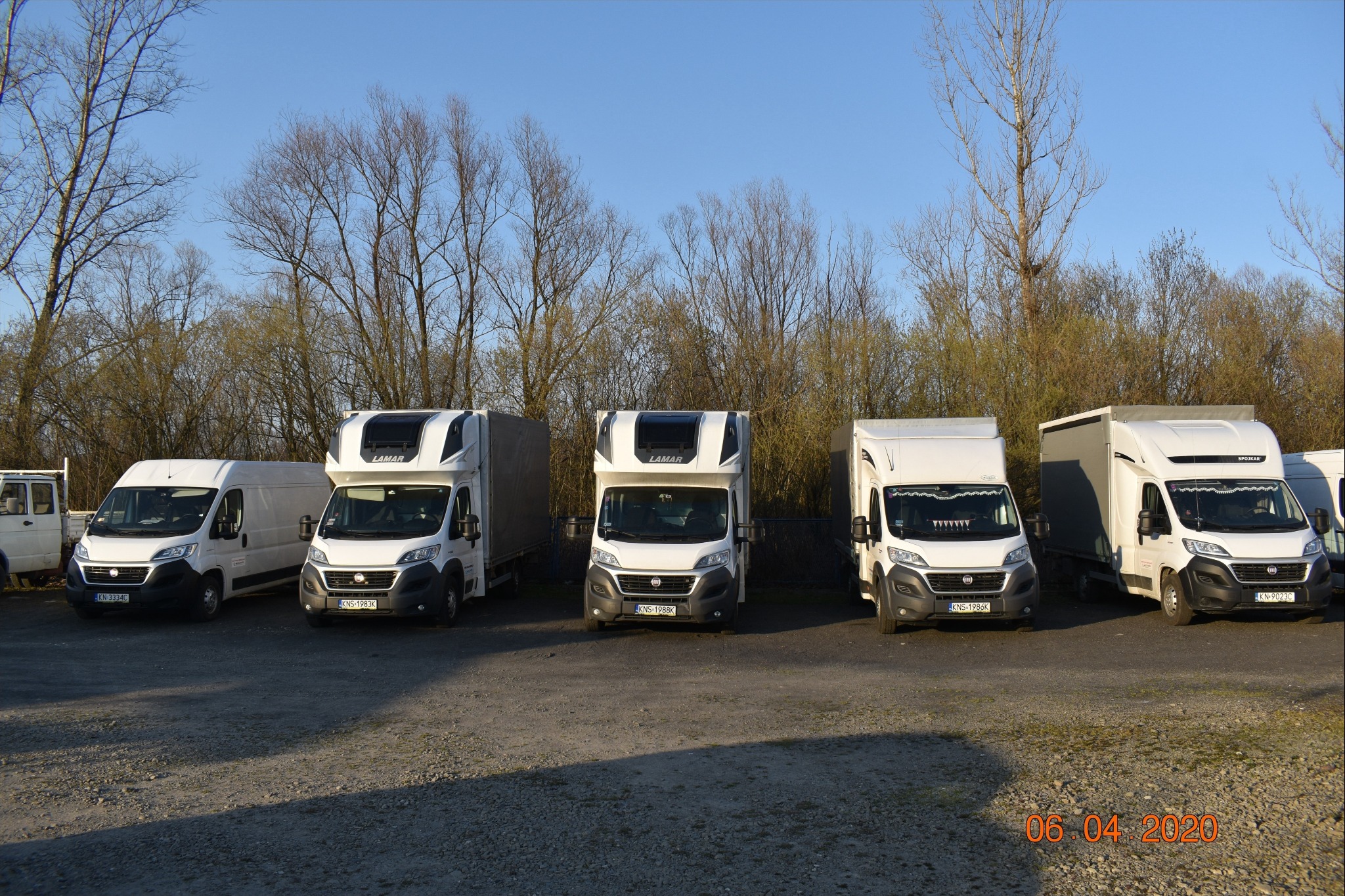 Flota białych busów transportowych firmy z Nowego Sącza, zaparkowana na żwirowym placu w słoneczny dzień, z widocznymi numerami rejestracyjnymi i nazwami firm LAMAR i SPOJKAR na niektórych pojazdach.