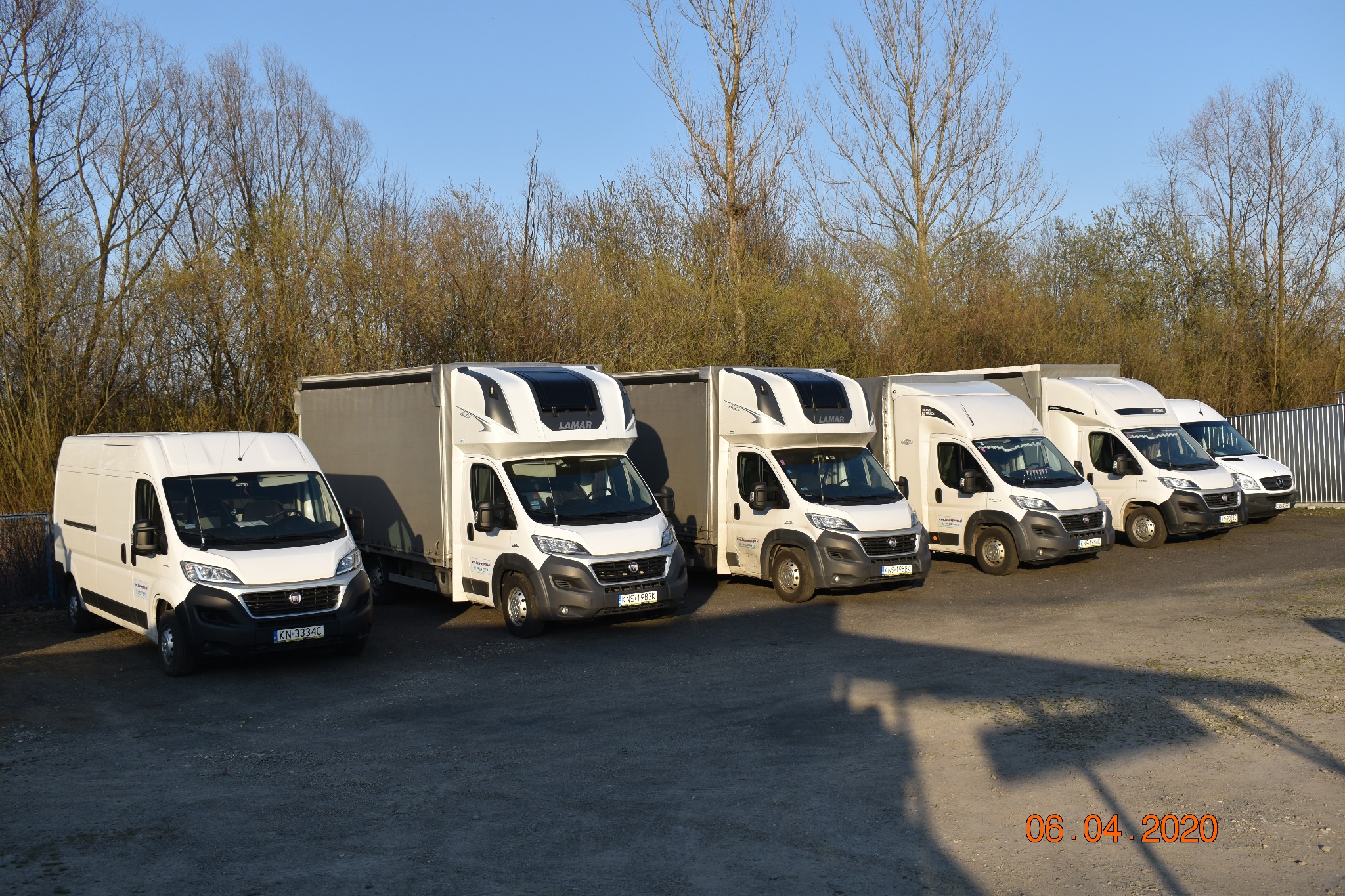 Flota białych busów dostawczych marki Fiat ustawionych w rzędzie na parkingu, z widocznymi numerami rejestracyjnymi i logotypami firm transportowych.