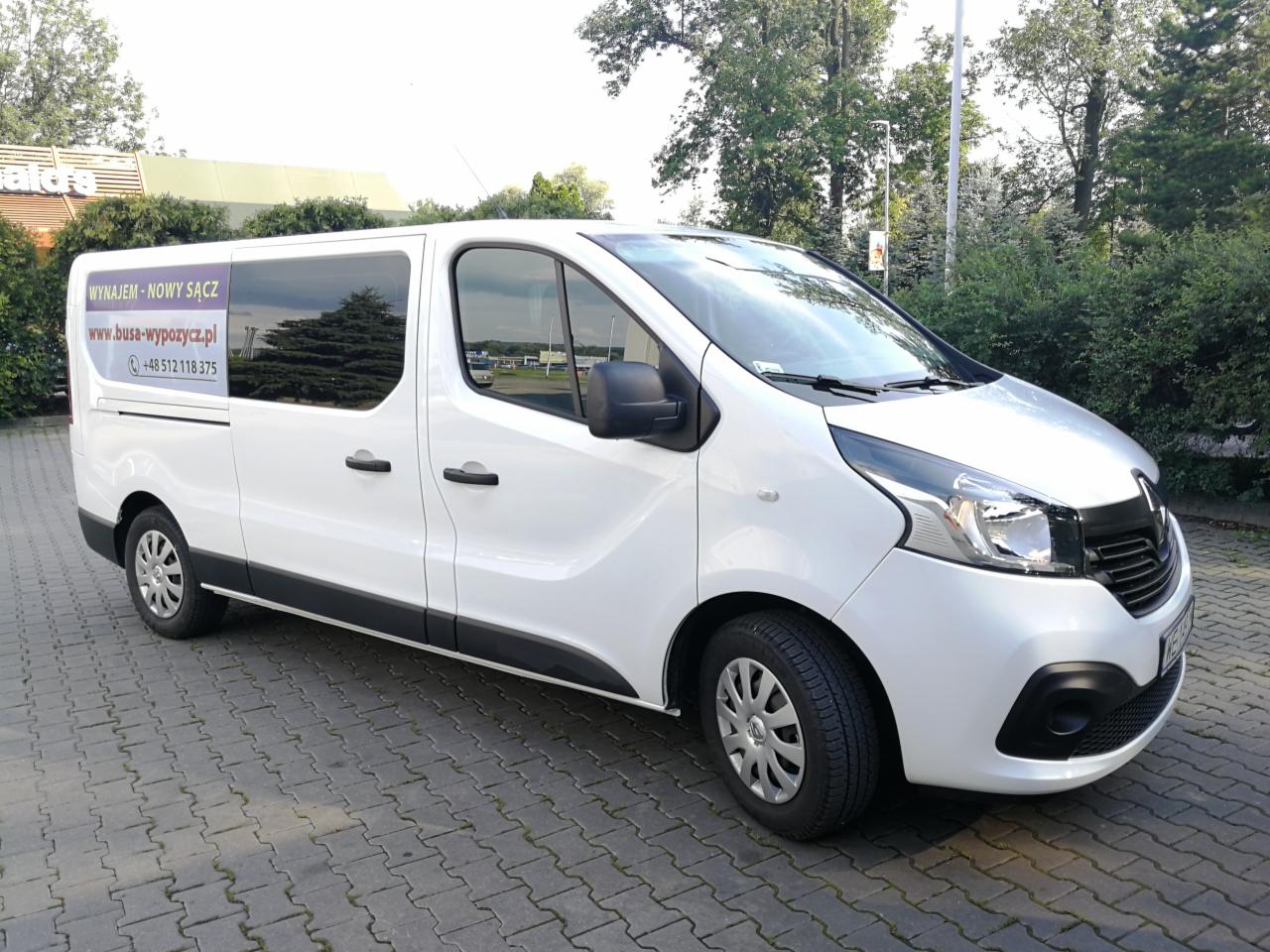 Biały Renault Trafic z przyciemnianymi szybami, zaparkowany na kostce brukowej przed zielenią, z naklejką reklamową 'Wynajem - Nowy Sącz' i adresem strony www na bocznej szybie.