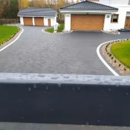 Elegancka posesja z ciemnoszarą kostką brukową ułożoną na podjeździe i chodniku, obrzeża wykończone białym pasem i drobnym żwirem, w tle dwa garaże z drewnianymi bramami i dom z białymi ścianami.