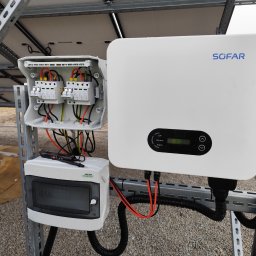 Instal Smart - Biały inwerter SOFAR z podłączonymi przewodami i skrzynką bezpiecznikową na metalowej konstrukcji, instalacja fotowoltaiczna w trakcie montażu, widoczne elementy pomiarowe.