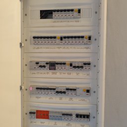 Instal Smart - Otwarta skrzynka elektryczna z bezpiecznikami i wyłącznikami różnicowoprądowymi, widoczne oznaczenia obwodów i zabezpieczeń. Instalacja wewnątrz budynku.