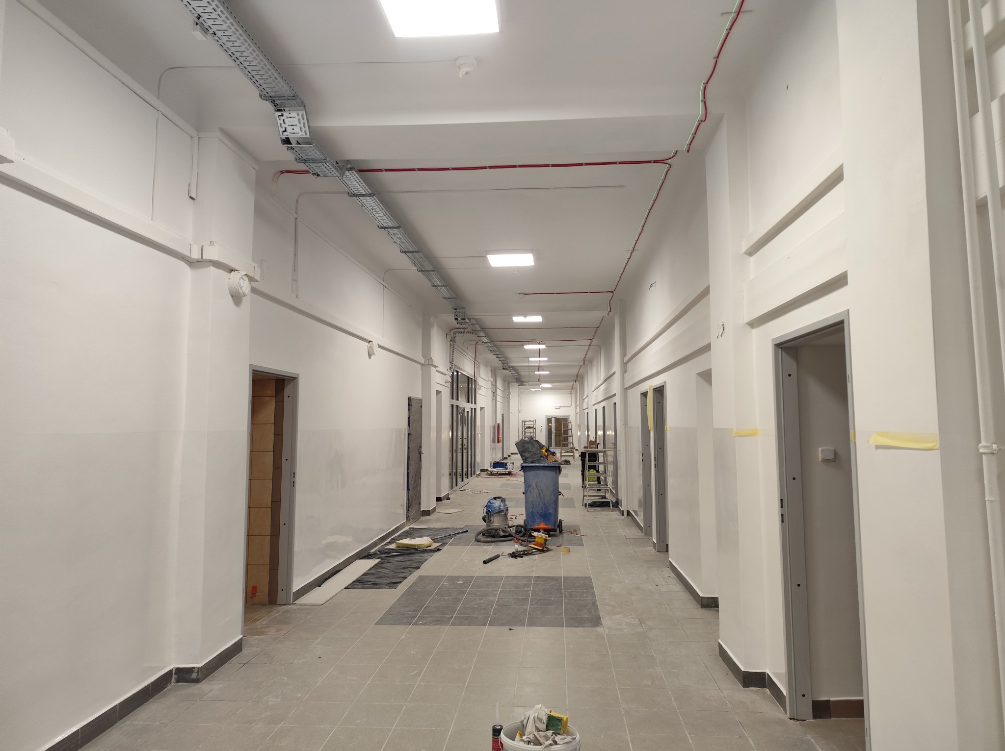 Długi korytarz w trakcie remontu: widoczne kable, rury pod sufitem, narzędzia i materiały budowlane. Szare płytki na podłodze, białe ściany.